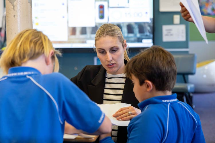 teacher kneeling down and assisting two students that are working at their desks.